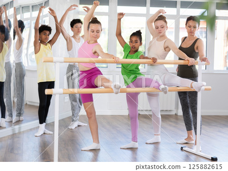 Teenage girls doing leg stretching exercise holding by ballet barre during dance courses 125882615
