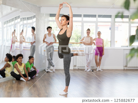 Gracious middle-aged female trainer demonstrating professional ballet poses to teenage boys and girls 125882677