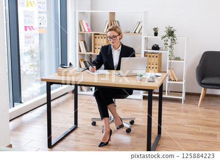 Professional woman in her thirties seated at desk in modern office analyzes documents and works on laptop. The workspace features natural light, organized shelves enhancing productivity and focus. 125884223