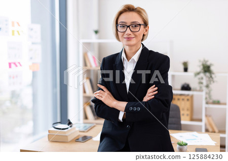 Professional adult female with confident look wearing formal suit in modern office. Business workspace with desk, plants, and shelves in background. Represents success, professionalism, motivation 125884230