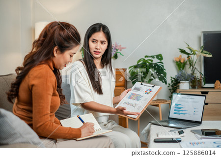 Two women discussing business strategies with charts and a laptop in a modern office environment. 125886014
