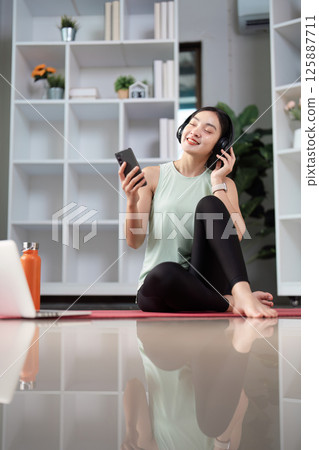 Happy Asian woman listening to music on headphones while using smartphone at home, embracing wellness and relaxation. 125887711