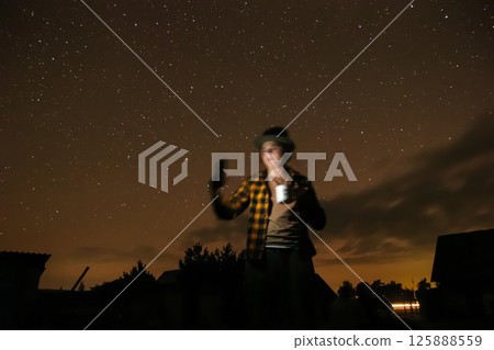 Night scene: woman gazing at the starry sky. Blurred lights and a phone in hand. Travel photography. Woman looking at the night sky full of stars. Blurred background with city lights 125888559
