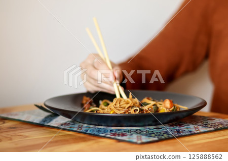 Woman eating noodles with chopsticks. Asian cuisine. Close-up of a woman using chopsticks to eat noodles. Delicious Asian food Woman eating noodles with chopsticks. Asian cuisine. Close-up of a woman using chopsticks to eat noodles. Delicious Asian food 125888562