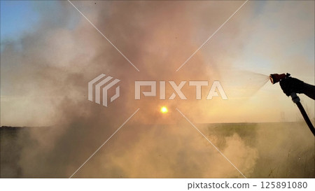 Extinguishing fires with improvised means in the yard. smoke from a fire in nature against a bright sunset. Yellow-gray smoke from the fire rises into the sky. 125891080