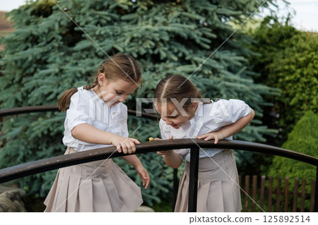 Twins in white outfits stand close together and lean toward the railing. A snail crawls on the wooden surface, attracting the girls' attention. 125892514