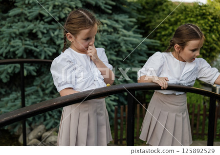 The twins observe the snail moving along the railing. Attentive girls in identical outfits create a sense of harmony. The twins observe the snail moving along the railing. Attentive girls in identical outfits create a sense of harmony. 125892529
