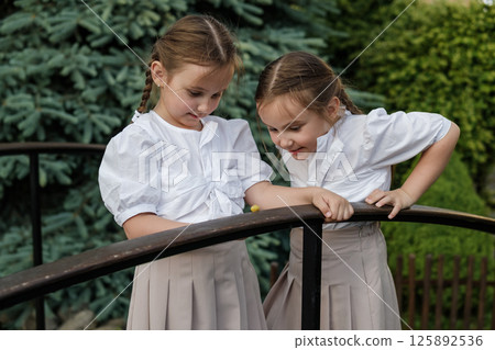 Little twins in light clothing lean over the snail. The girls stand near the railing, observing nature. Little twins in light clothing lean over the snail. The girls stand near the railing, observing nature. 125892536