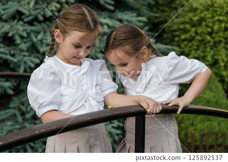 Little twins in light clothing lean over the snail. The girls stand near the railing, observing nature. 125892537