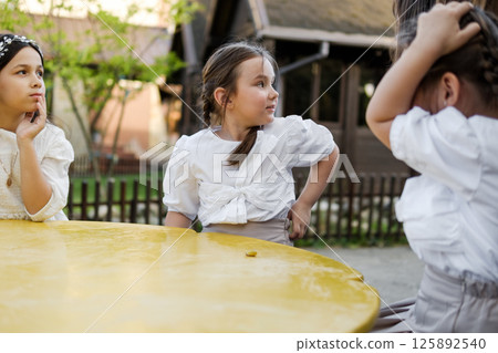 Cute girls are sitting at a children's table. The twin girls are dressed in white blouses, and their older sister is sitting next to them in a delicate dress. 125892540