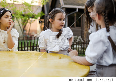 Cute girls are sitting at a children's table. The twin girls are dressed in white blouses, and their older sister is sitting next to them in a delicate dress. 125892541