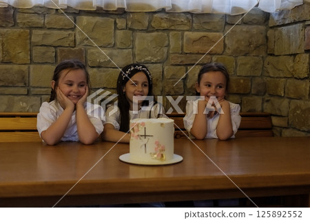 Three girls create a warm atmosphere near the brightly decorated cake. The twin girls with matching hairstyles sit close together, admiring the decorations. Three girls create a warm atmosphere near the brightly decorated cake. The twin girls with matching hairstyles sit close together, admiring the decorations. 125892552
