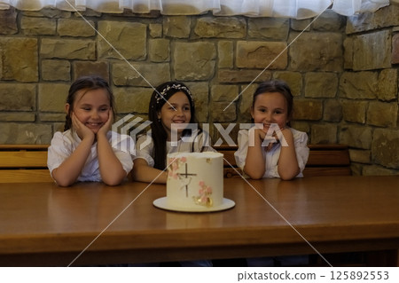 Three girls create a warm atmosphere near the brightly decorated cake. The twin girls with matching hairstyles sit close together, admiring the decorations. 125892553