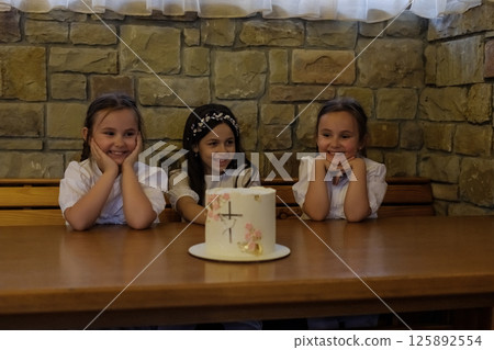 The twin girls and their older sister settled at the wooden table in a festive setting. The decorated cake became the focal point for all the girls. The twin girls and their older sister settled at the wooden table in a festive setting. The decorated cake became the focal point for all the girls. 125892554