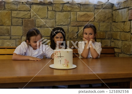The girls in white clothing sit at the table near a delicious-looking cake. The twin girls with braids look touching beside the older girl. The girls in white clothing sit at the table near a delicious-looking cake. The twin girls with braids look touching beside the older girl. 125892556