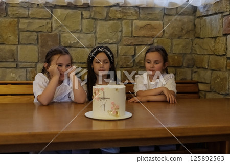 The twin girls with braids and the girl in a dress sit near the festive dessert. The girls appear focused and enchanted. The twin girls with braids and the girl in a dress sit near the festive dessert. The girls appear focused and enchanted. 125892563