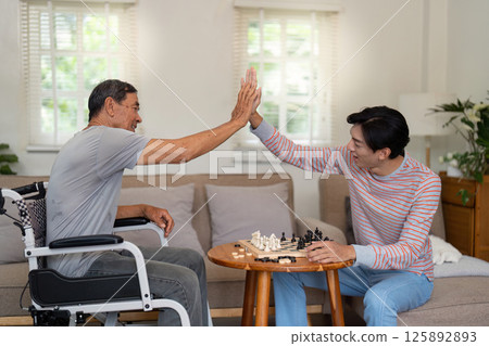 Father and son celebrating a chess move with a high five in a cozy living room, emphasizing family joy and connection. 125892893