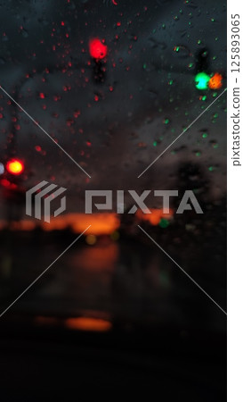 Blurred view through rain-soaked windshield capturing traffic lights glowing against dark stormy sky with hints of sunset creating dramatic urban atmosphere Blurred view through rain-soaked windshield capturing traffic lights glowing against dark stormy sky with hints of sunset creating dramatic urban atmosphere 125893065