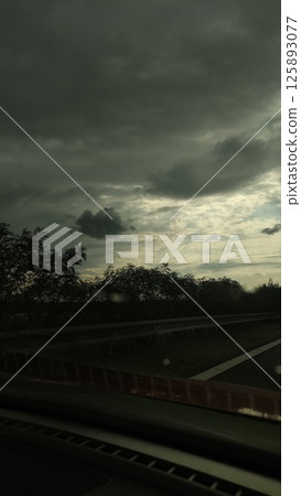 Dramatic overcast sky with swirling clouds and hints of sunlight breaking through, creating a moody atmosphere along a winding roadside landscape 125893077
