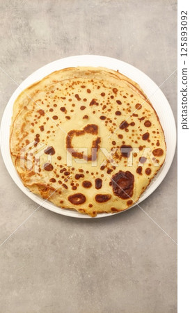Golden-brown pancake with unique heart-shaped pattern on surface, served on white plate against textured gray background, perfect for breakfast or brunch culinary delight Golden-brown pancake with unique heart-shaped pattern on surface, served on white plate against textured gray background, perfect for breakfast or brunch culinary delight 125893092