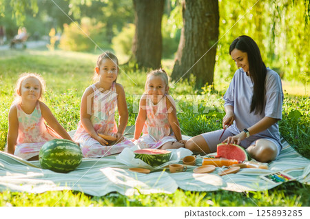 Cute little girls of 4-5 years old and their mother are eating a delicious watermelon against the background of green nature. Healthy lifestyle. Cute little girls of 4-5 years old and their mother are eating a delicious watermelon against the background of green nature. Healthy lifestyle. 125893285
