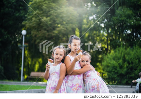 Three sisters stand side by side in a park enjoying an ice cream. 125893339