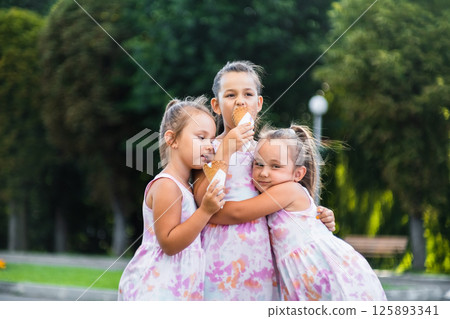 Three sisters, dressed in identical dresses, stand holding hands and enjoying ice cream. Three sisters, dressed in identical dresses, stand holding hands and enjoying ice cream. 125893341