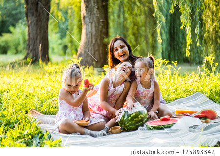 The girls and their mother are eating watermelon, their dresses are a little dirty. The twins do not pay attention, laughing together.  125893342