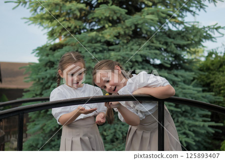 Schoolgirls in identical outfits look like an illustration from a children's book. The snail on the railing becomes the center of their world for a moment. Schoolgirls in identical outfits look like an illustration from a children's book. The snail on the railing becomes the center of their world for a moment. 125893407