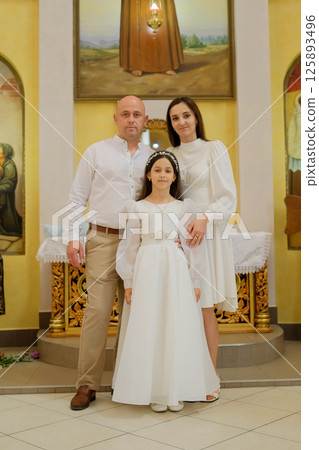 Inside the church, a young family gathers to celebrate a spiritual ceremony. A little girl in a cream dress is surrounded by caring parents. 125893496