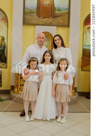 A family couple stands with three daughters in the church. The First Communion of a sweet girl. A family couple stands with three daughters in the church. The First Communion of a sweet girl. 125893497