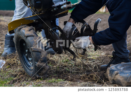 Preparing the land for planting with a walk-behind tractor. A farmer installs blades on a walk-behind tractor, preparing to plow well-weeded soil, preparing the land for the upcoming planting season. Preparing the land for planting with a walk-behind tractor. A farmer installs blades on a walk-behind tractor, preparing to plow well-weeded soil, preparing the land for the upcoming planting season. 125893788
