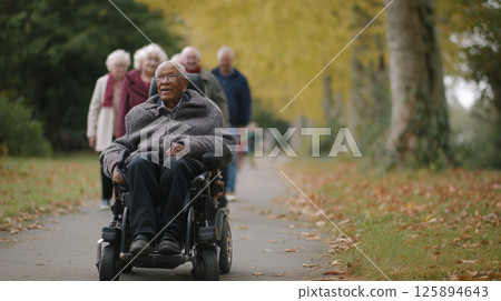 An elderly man with a warm smile sits in a wheelchair on a pathway in a park, enjoying the fresh air as friends walk alongside him amidst vibrant autumn foliage. 125894643