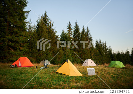 Solar panel in grassy field with tents in background highlights eco-friendly camping setup, surrounded by dense evergreen trees in evening. Integration of renewable energy in outdoor activities. 125894751