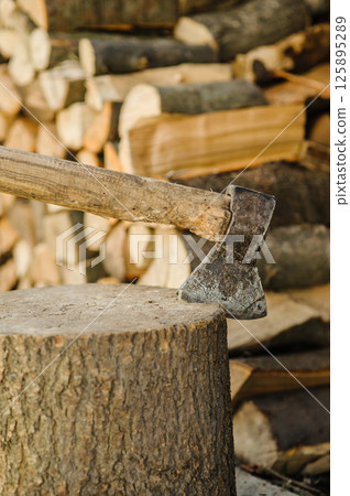 An axe is stuck in a sturdy stump. Behind it, there are many chopped and stacked logs. An axe is stuck in a sturdy stump. Behind it, there are many chopped and stacked logs. 125895289