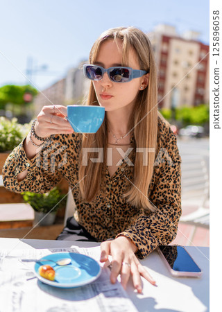 Young woman wearing stylish sunglasses, savoring a cup of coffee and a delightful pastry at an outdoor cafe. Newspaper and smartphone resting on the table, capturing a serene morning moment 125896058