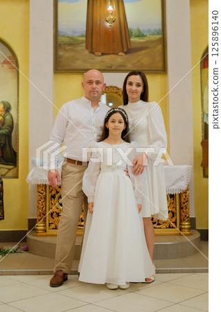 Inside the church, a young family gathers to celebrate a spiritual ceremony. A little girl in a cream dress is surrounded by caring parents. 125896140