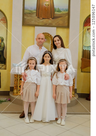 A festive family stands in a brightly lit church. The girls in elegant outfits form a tender family circle near the parents. A festive family stands in a brightly lit church. The girls in elegant outfits form a tender family circle near the parents. 125896147