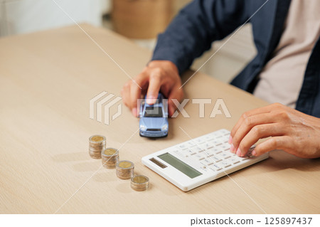 A person uses a calculator near coins and a car model, calculating auto loan payments and taxes. Perfect for car sales, finance, insurance, and banking concepts in financial planning. A person uses a calculator near coins and a car model, calculating auto loan payments and taxes. Perfect for car sales, finance, insurance, and banking concepts in financial planning. 125897437
