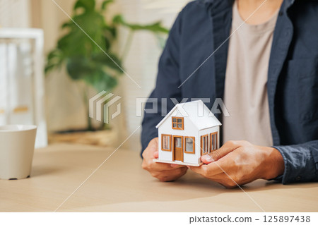 Close-up of a real estate agent hand holding a small house model, symbolizing property, home loan, and finance concepts. Represents the role of an agent in real estate, housing, and loan planning. Close-up of a real estate agent hand holding a small house model, symbolizing property, home loan, and finance concepts. Represents the role of an agent in real estate, housing, and loan planning. 125897438
