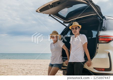 Asian couple is posing in back of a car enjoying beautiful views on the ocean beach. lifestyle man and the woman is wearing hat. Scene is relaxed and carefree, love and travel summer trip concept 125897447