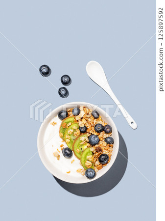 Natural yogurt with granola, kiwi and blueberries in a bowl on a blue background with berries. Natural yogurt with granola, kiwi and blueberries in a bowl on a blue background with berries. 125897592
