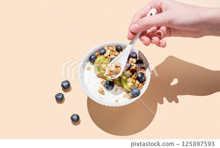 Hand holding spoon natural yogurt with granola, kiwi and blueberry in bowl on beige background Hand holding spoon natural yogurt with granola, kiwi and blueberry in bowl on beige background 125897593