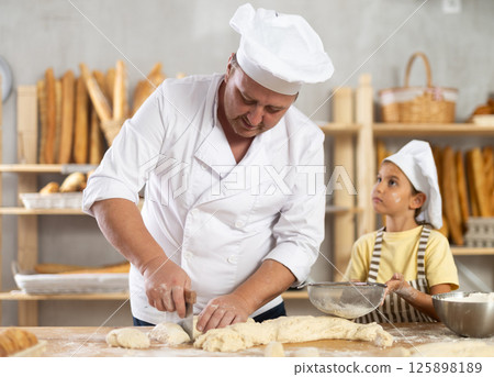 Baker man cuts raw dough with knife. Little daughter helps him by sprinkling flour over dough through sieve Baker man cuts raw dough with knife. Little daughter helps him by sprinkling flour over dough through sieve 125898189