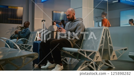 Airport Terminal: Man with Suitcase Sitting in Departure Lounge After Security Check, Using Phone Airport Terminal: Man with Suitcase Sitting in Departure Lounge After Security Check, Using Phone 125898282