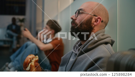 Airport Terminal: Close Up Shot of Man Eating Burger, Sitting on the Floor in Departure Lounge 125898284