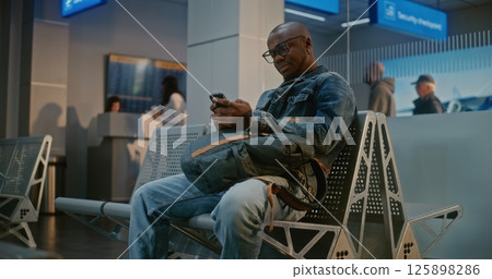 Airport Terminal: Man with Backpack Sitting in Boarding Lounge After Security Check, Using Phone Airport Terminal: Man with Backpack Sitting in Boarding Lounge After Security Check, Using Phone 125898286