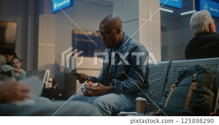 Man Eating Fast Food and Using Smartphone Sitting in Departure Lounge of Airline Hub 125898290