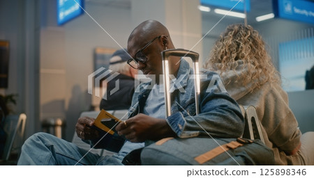 Man with Luggage Sitting and Waiting for Flight, Using Smartphone in Departure Lounge of Crowded Airline Hub Man with Luggage Sitting and Waiting for Flight, Using Smartphone in Departure Lounge of Crowded Airline Hub 125898346