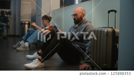 Airport Terminal: Man Scrolling Smartphone, Sitting on the Floor, Waiting for Delayed Flight 125898353